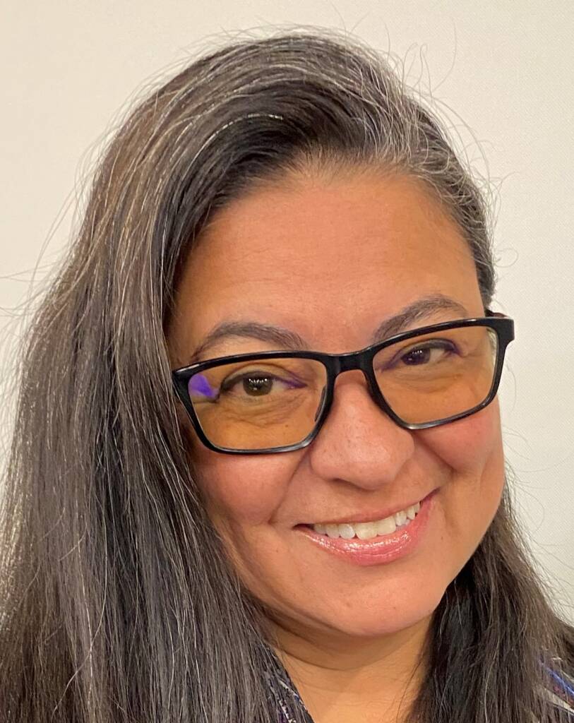 A head to shoulder photo of a Hispanic female smiling with black glasses, long brown hair, tan skin, and brown eyes.