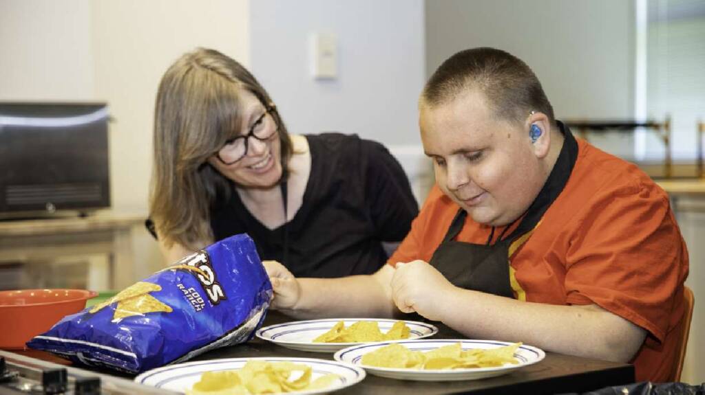 A DeafBlind man wears a hearing aid and holds a bag of chips next to his instructor
