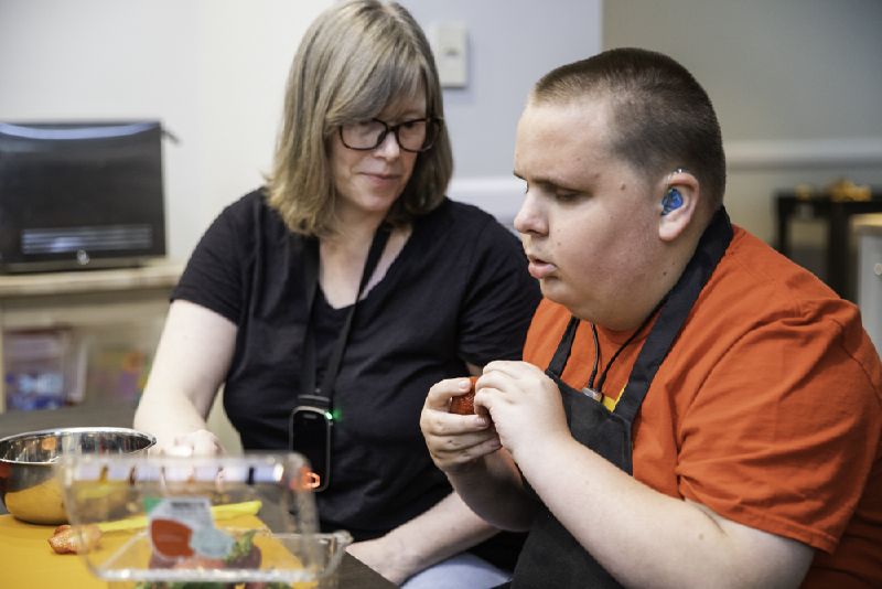 A DeafBlind man wears a hearing aid and prepares to cut strawberries next to his instructor