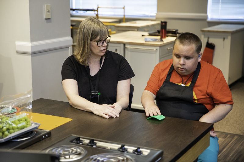 A DeafBlind man wears a hearing aid and touches a measuring cup to learn Braille alongside his instructor