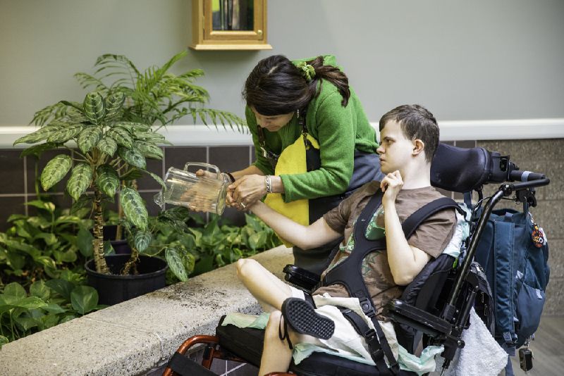 A DeafBlind man sits in an adaptive wheelchair and touches the bottom of a pitcher in a garden with the assistance of his instructor
