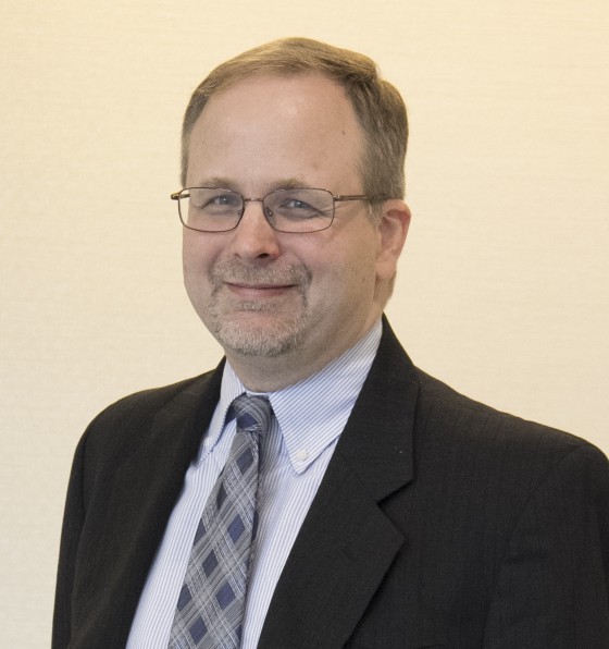 A man named Jeff Kunkel wearing a suit, tie, and glasses smiles into the camera