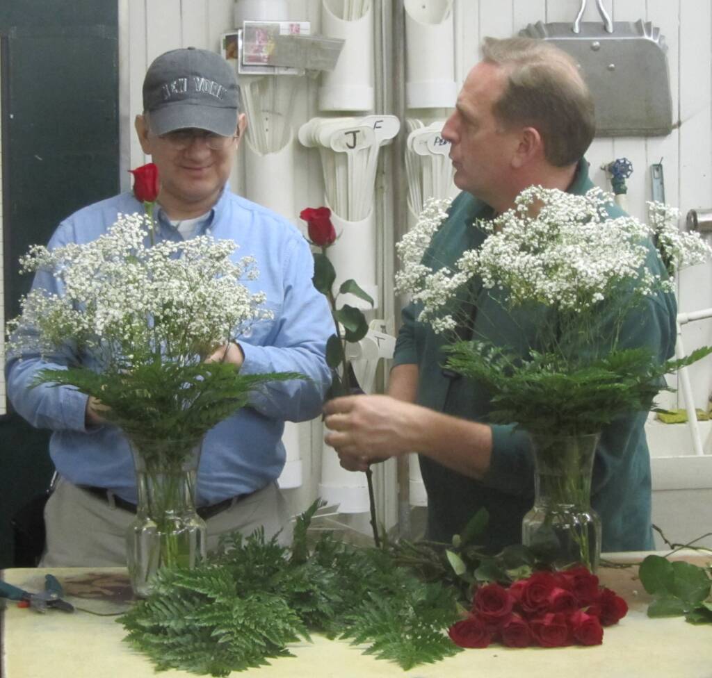 A man arranging roses and baby's breath in a vase at a florist shop while a man hands him a rose