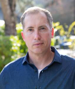 A middle-aged smiling white male with salt and pepper hair wearing a navy-blue collared shirt in front of a tree and bushes 