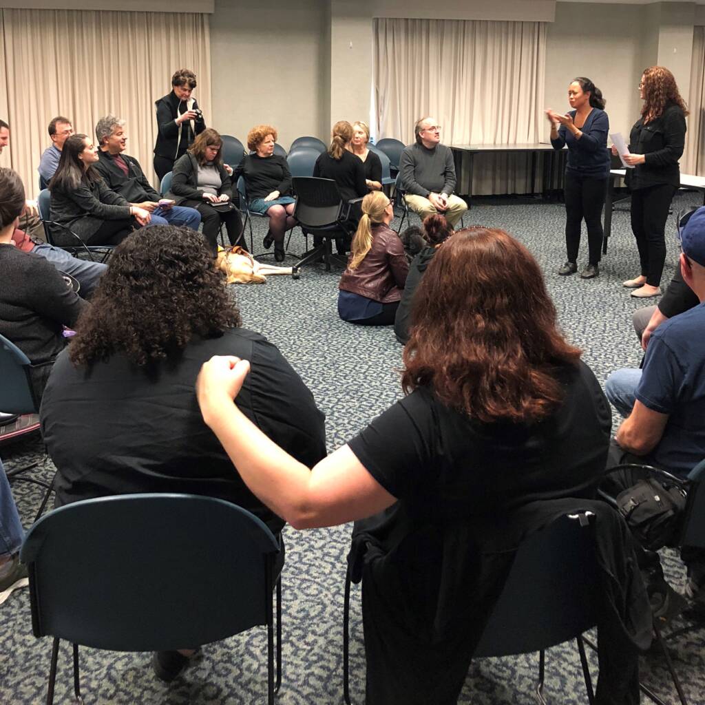 A woman practicing haptics by touching another woman's back with her hand. They sit in a circle with other people and look at a presenter.