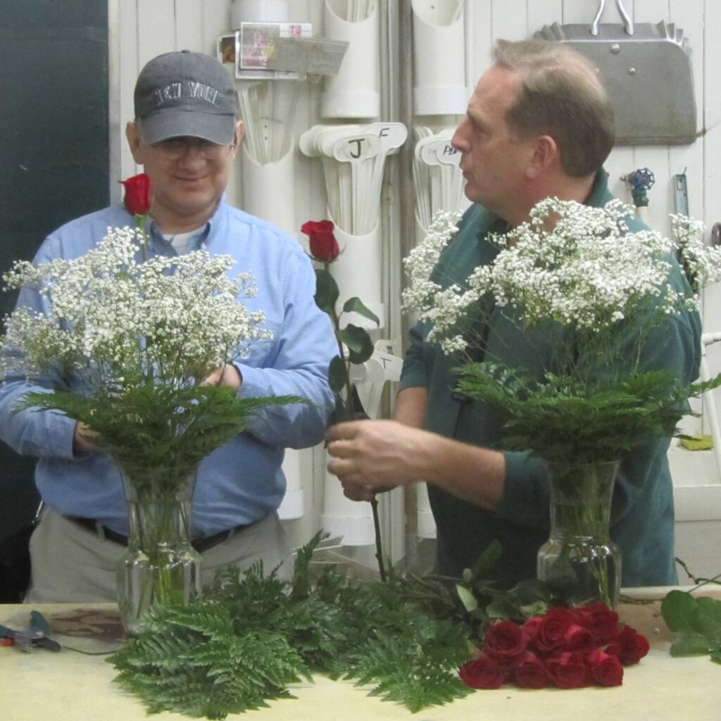 A man arranging roses and baby's breath in a vase at a florist shop while a man hands him a rose