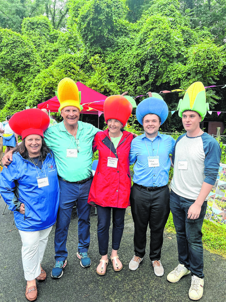 A group of people standing together and smiling outside. They wear large hats that look like corn, apples, and berries.