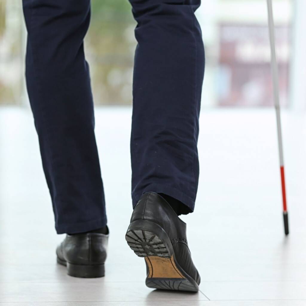 A close up shot of someone standing outside and holding a white cane. In the photo, only the feet, back of legs, and bottom half of white cane are showing.
