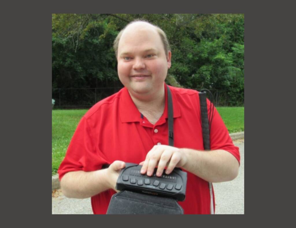 Scott Davert smiling and holding a braille display and white cane outside