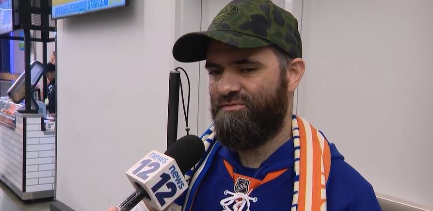 A man named Stuart Campbell talking into a News 12 microphone. He is holding a white cane and wearing an Islanders jersey.
