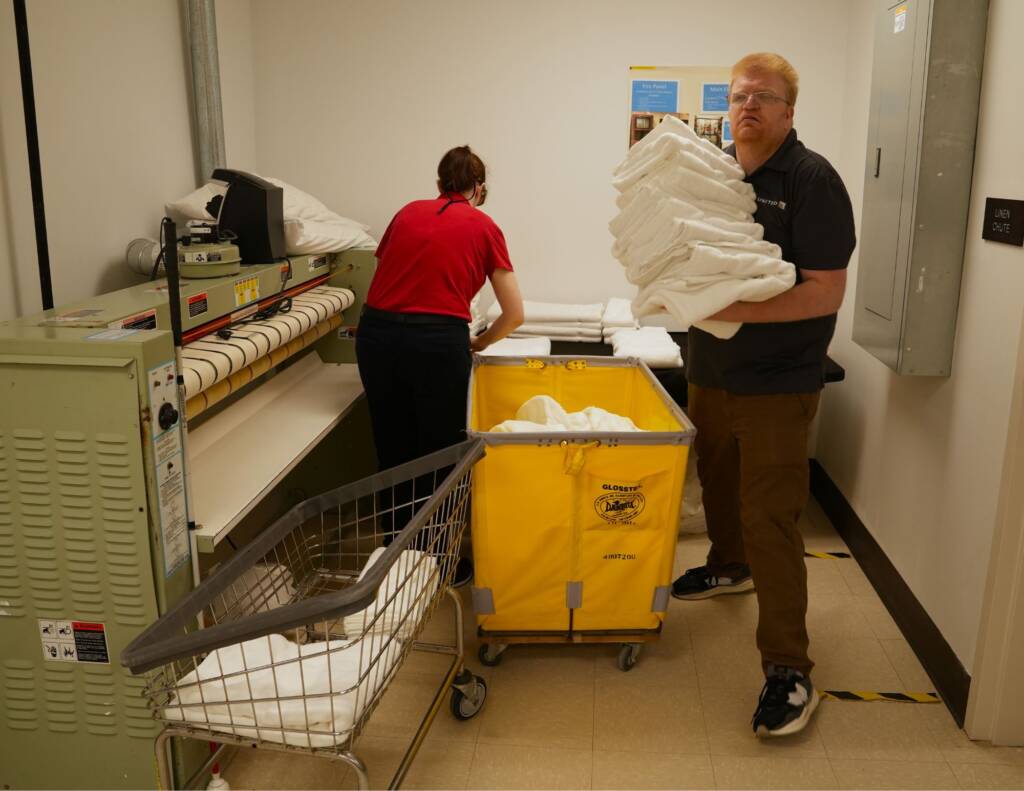 a man holding a large pile of folded white towels next to laundry crates
