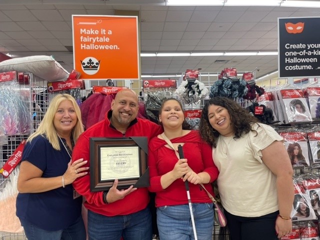 Ms. Farah standing with 3 people inside Savers. The man next to her holds a framed employee recognition award. Ms. Farah holds a white cane.