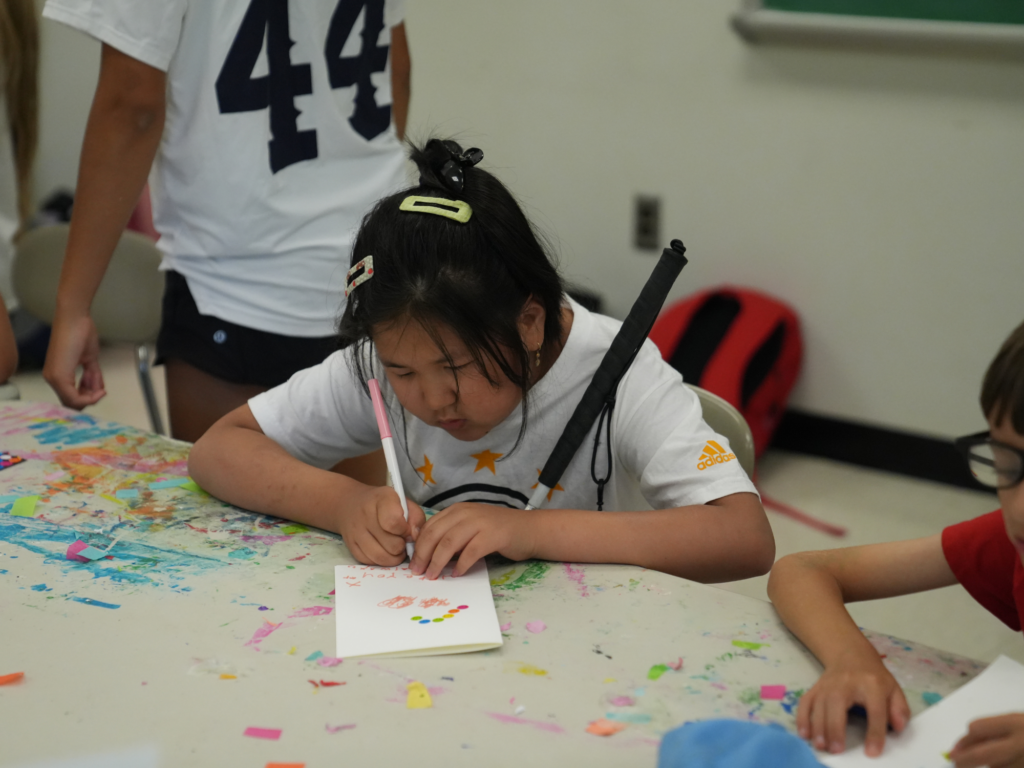 A young girl writing and drawing on a piece of paper with a pink marker and holding a white cane