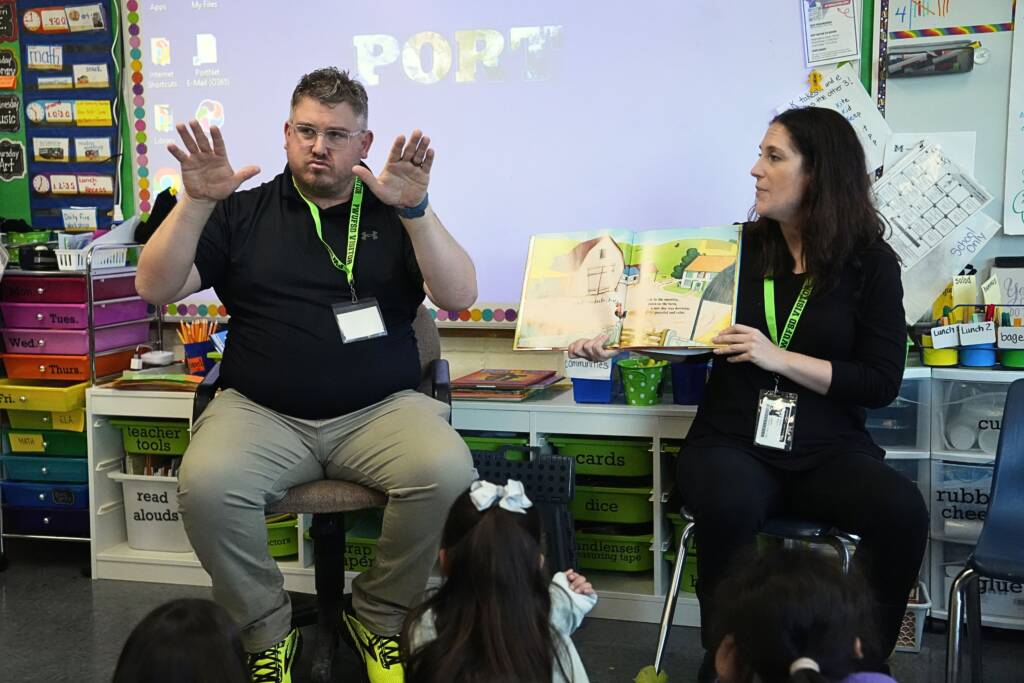 a man signing in a classroom next to a woman holding open a picture book
