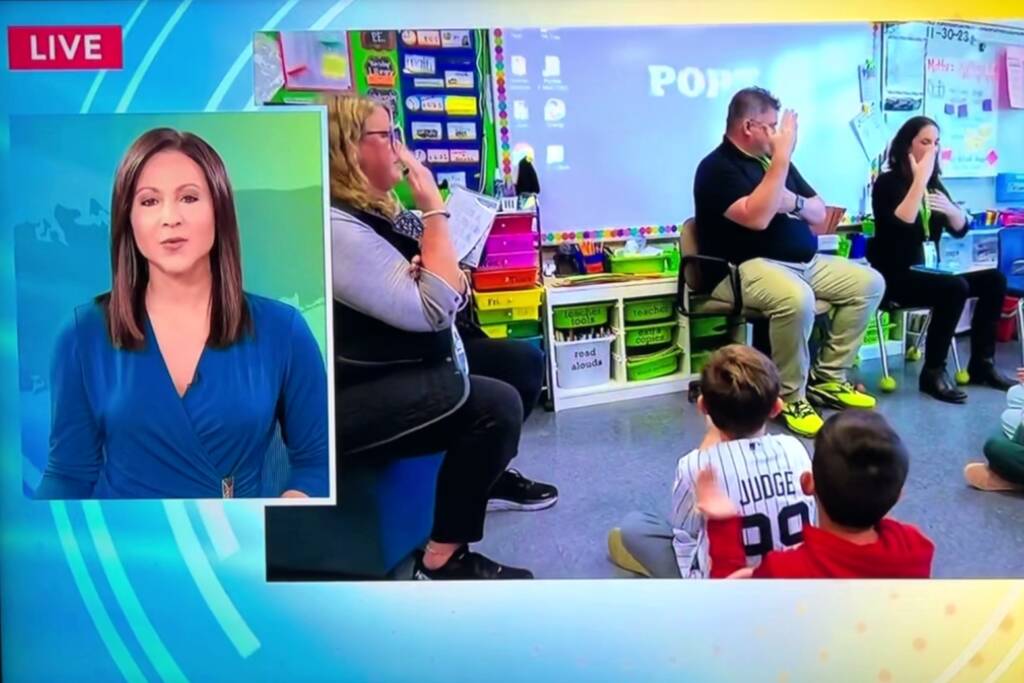 A newscaster looking into the camera, three adults doing sign language, and children sitting on the floor watching the adults