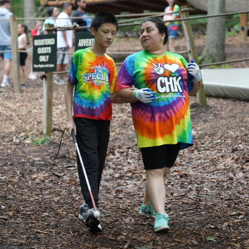 A boy holds a white cane and the arm of a woman outside. Both wear colorful Camp Helen Keller t shirts.