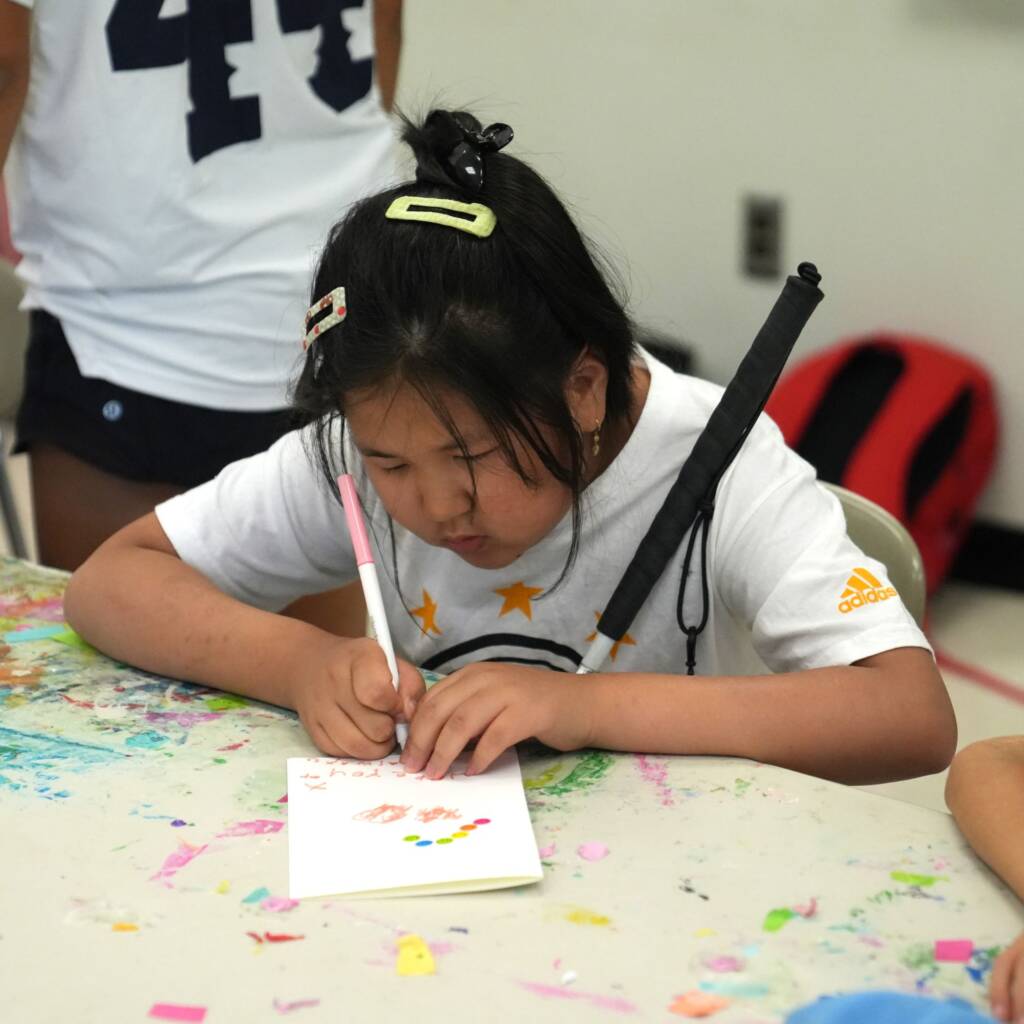 A girl writes on a paper card with a pink marker and holds a white cane