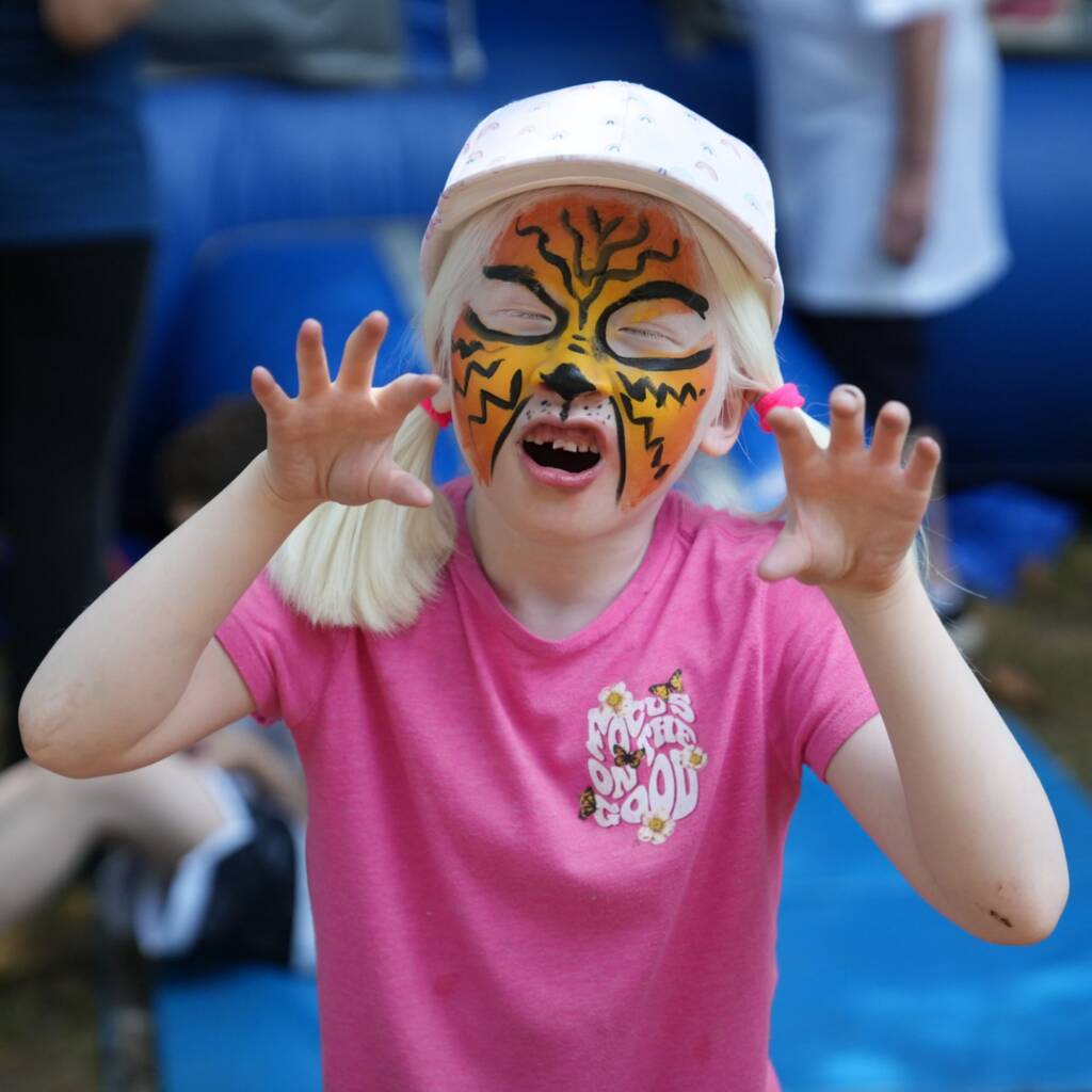 A girl with tiger face makeup makes a funny face and has her hands raised in a claw position.