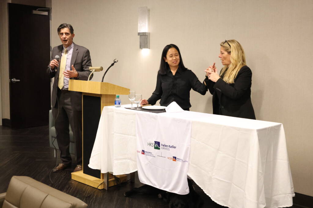 Two women communicate in tactile sign language as a man standing next to a podium speaks into a microphone. 