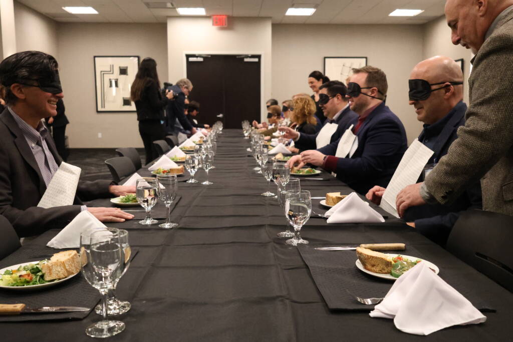 People sit at a long table wearing blindfolds. There are plates of food, drinking glasses, and cloth napkins on the table.