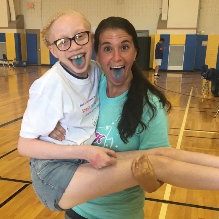 A woman named Stephanie MacLennan, who is the camp director, holding a girl camper in her arms inside a gymnasium. Both of them are smiling with their tongues sticking out. Both of their tongues are tinted blue.