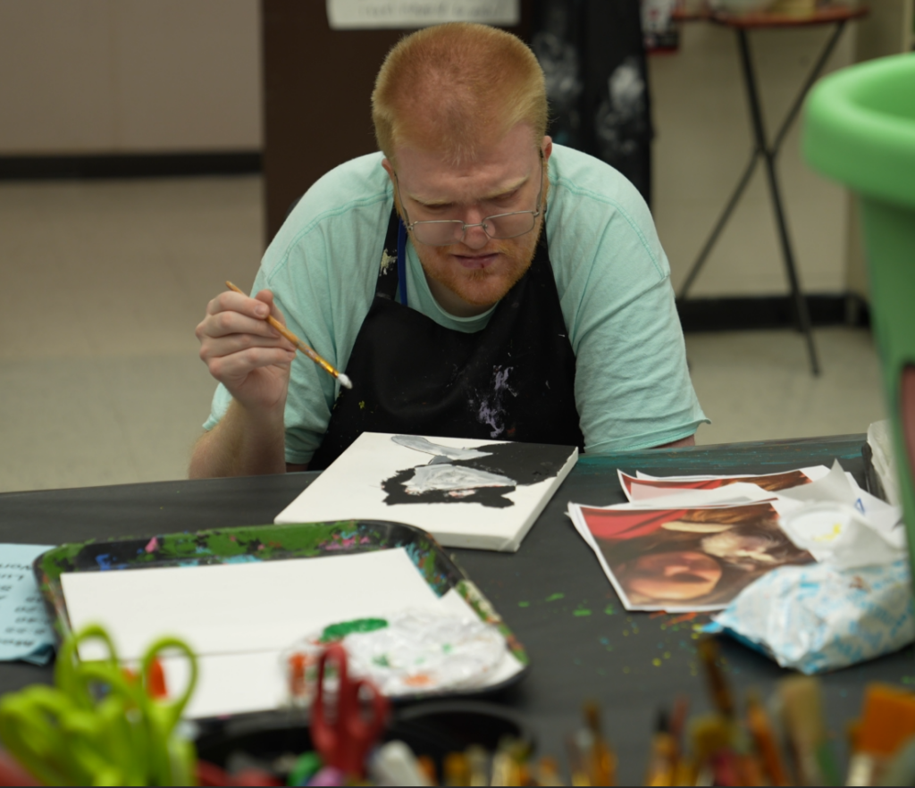 A man painting in an art classroom surrounded by art supplies