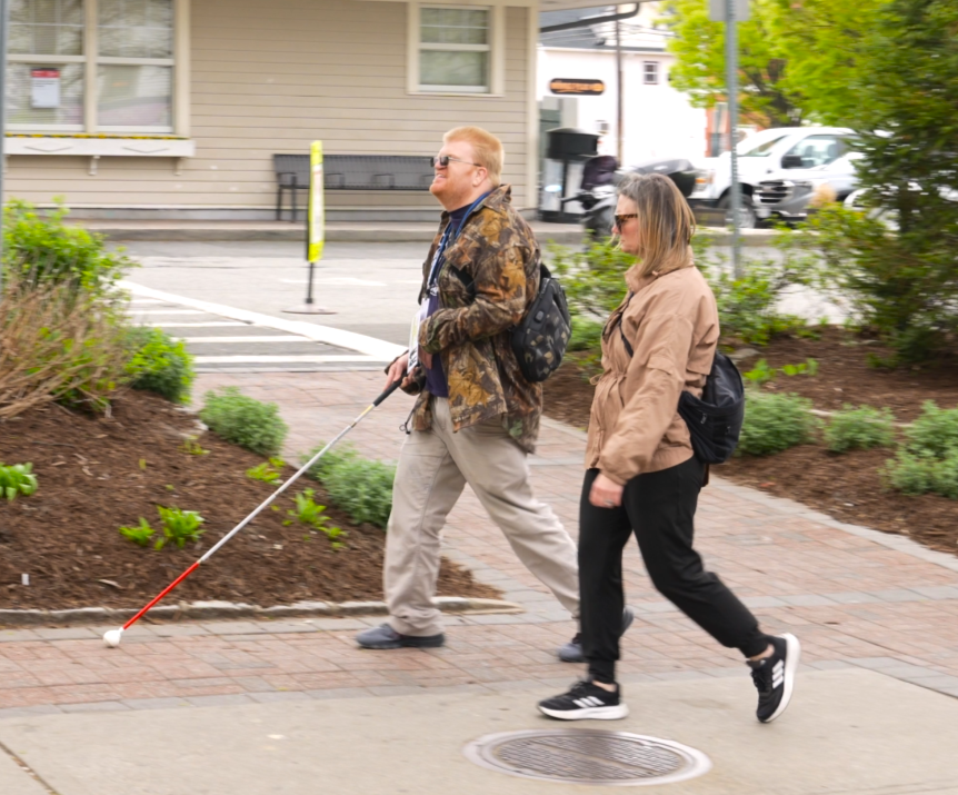 A man walking on a sidewalk with a white cane next to a woman