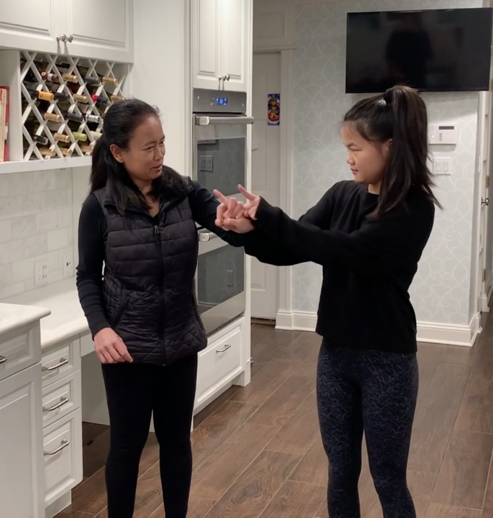 Two women are standing in a kitchen. The woman on the left has her hand on the other woman’s hand, tracking. The woman on the right is signing “go”.