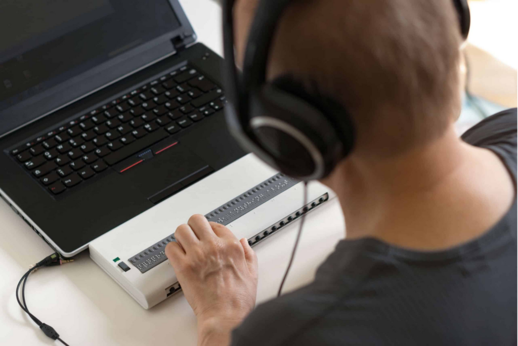 A man wearing headphones uses a laptop and braille display.  