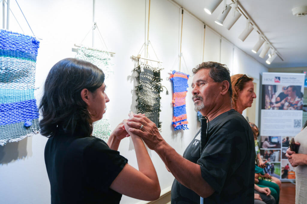 A man and woman communicate using tactile ASL to discuss the woven art pieces.