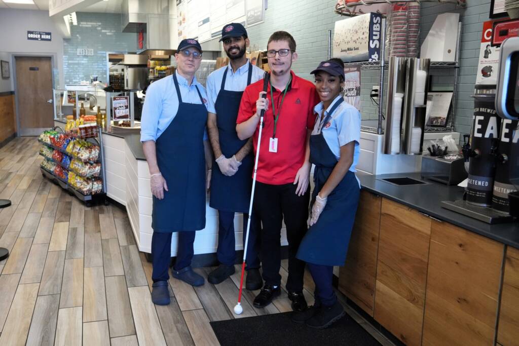 Jonathan, holding a white cane, stands next to 3 other employees of Jersey Mike’s for a group photo.