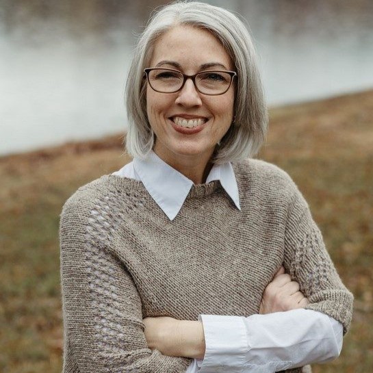 A Caucasian woman with platinum, chin-length hair and glasses is smiling directly at the camera. She is wearing a white blouse with a tan parka sweater over it. She is standing outside on a grassy hillside with a view of a lake behind her.