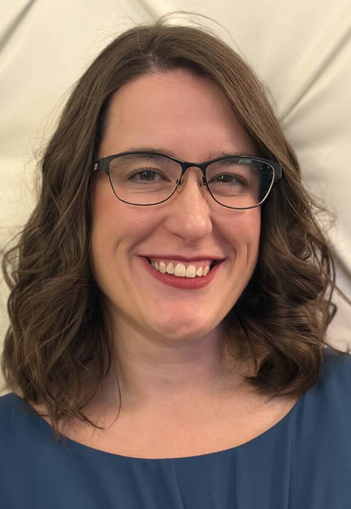 Headshot of Jenna Rydl, she is smiling with shoulder-length brown hair and black glasses.