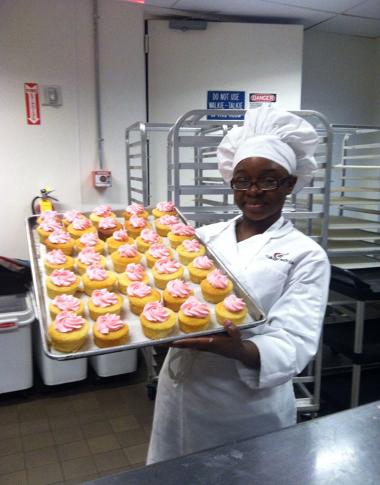 A girl in her early 20s is standing in a cafeteria kitchen. She is wearing glasses, a white chef's coat and a white chef's hat. She is smiling and holding a tray of cupcakes with pink frosting on them.