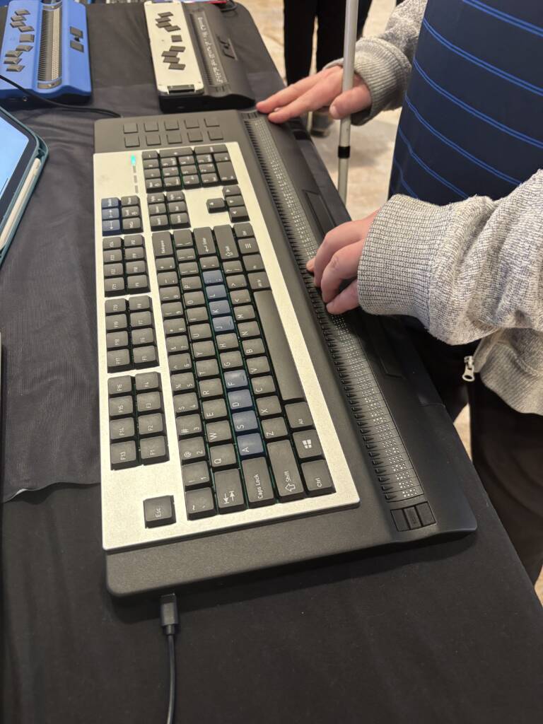 An Activator Pro braille display keyboard with a standard QWERTY layout and refreshable braille line along the bottom, sitting on a table with a person's hands positioned on it.