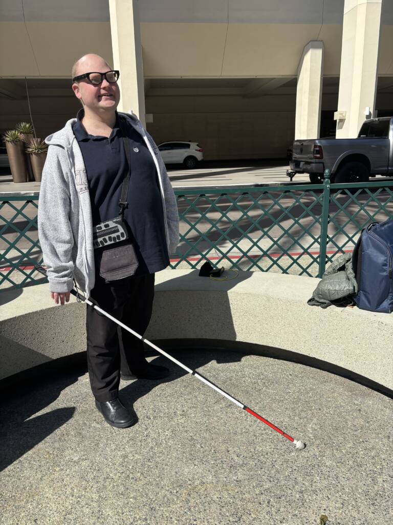 Scott standing outdoors holding a white cane with a red tip, wearing EchoVision smart glasses and carrying a braille display in a crossbody pouch.