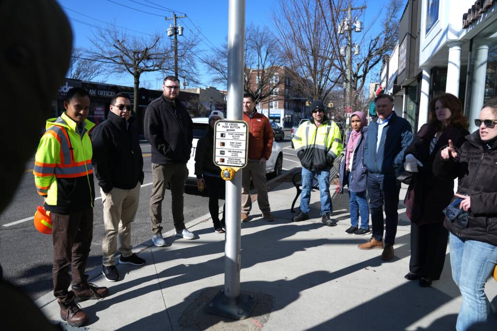A large group of people gathered at a street crossing. Several wear high-visibility safety vests, including traffic engineers from Nassau, Suffolk, and New York State. The group is positioned at an intersection with a pedestrian signal button visible on a pole.