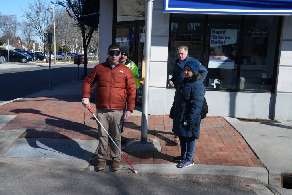 A man wearing a blindfold and holding a white cane stands on a sidewalk near a pedestrian crossing pole while others look on.