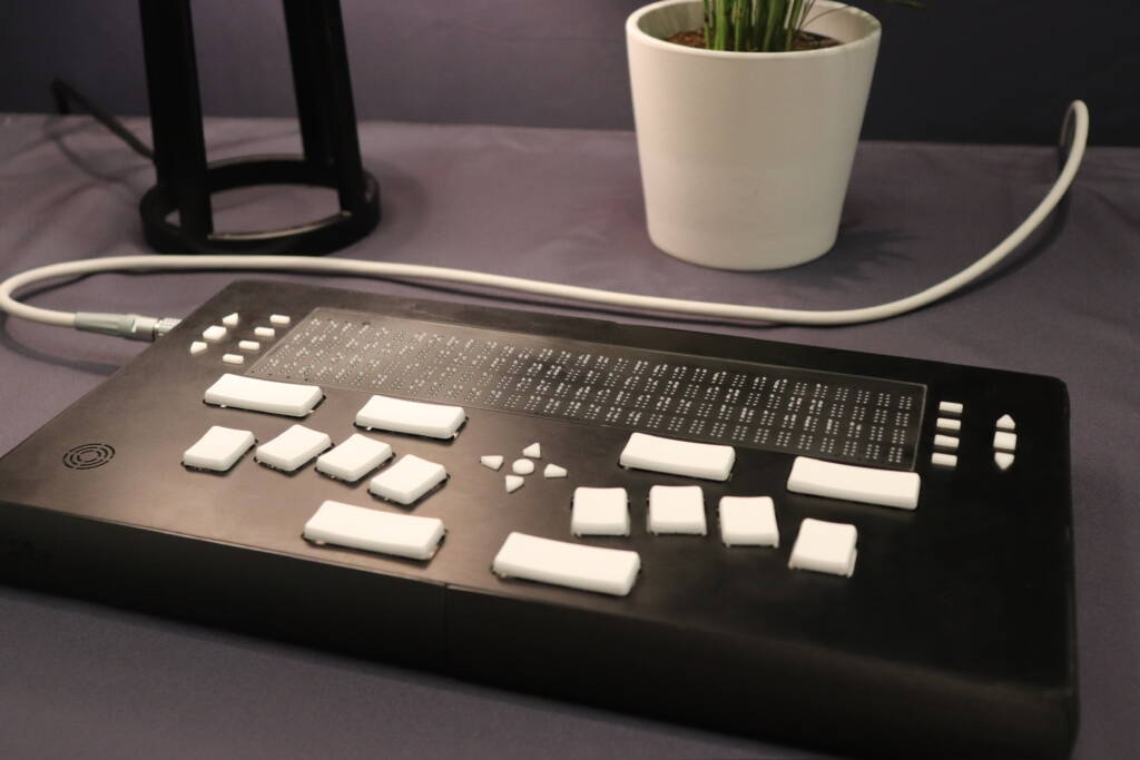 A black braille keyboard device with white keys and a braille display, called the Codex from NewHaptics, sitting on a surface with a small potted plant in the background.