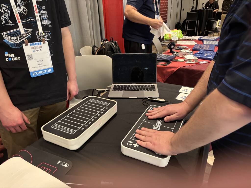 A trade show exhibit featuring assistive technology devices from OneCourt shaped like football fields, with a person demonstrating the "Kings" controller while an exhibitor with a badge stands nearby next to a laptop and promotional materials.