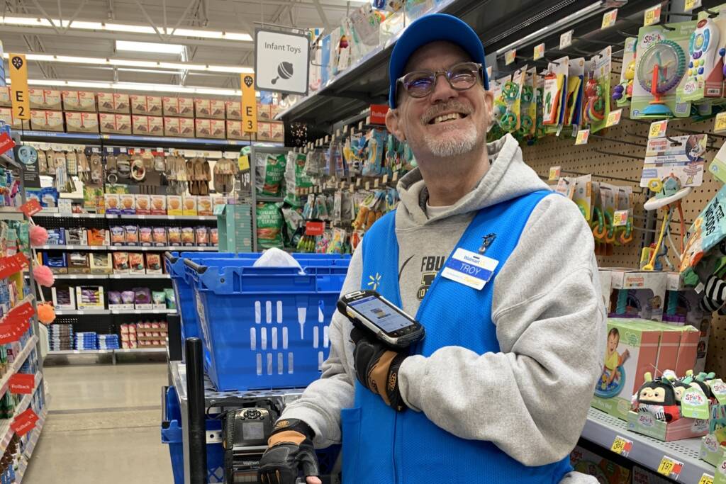 Troy stands smiling in front of a shopping cart in Walmart while holding a product scanning device.