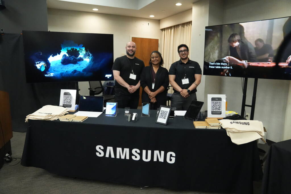 Representatives from Samsung stand together behind their vendor table alongside 2 large TV displays.