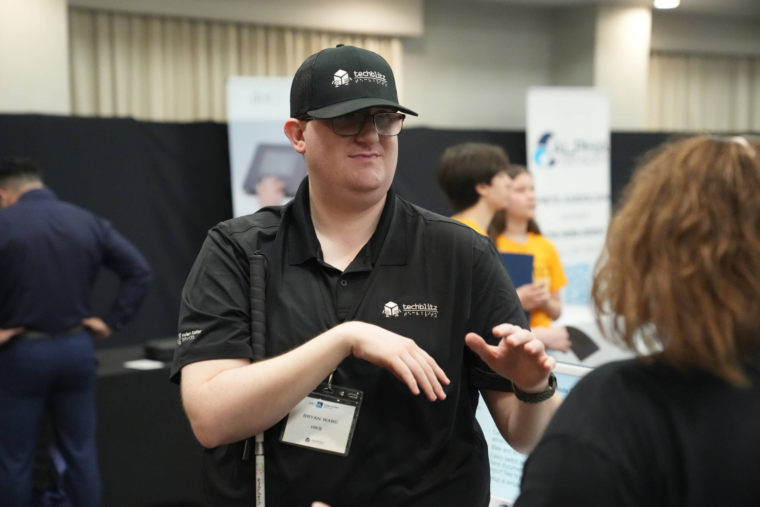 Bryan Ward is holding a white cane in his arm while signing in ASL. he is wearing a black Tech Blitz hat and shirt.
