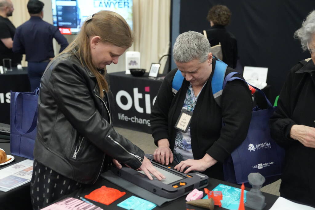 Chancey shows an attendee the Monarch braille display at the Andrew Heiskell Braille and Talking Book Library table.