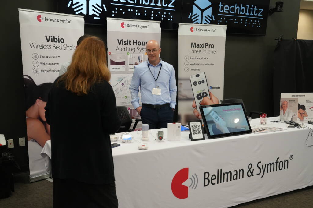 A man stands behind the Bellman & Symfon vendor table featuring products and devices on display.