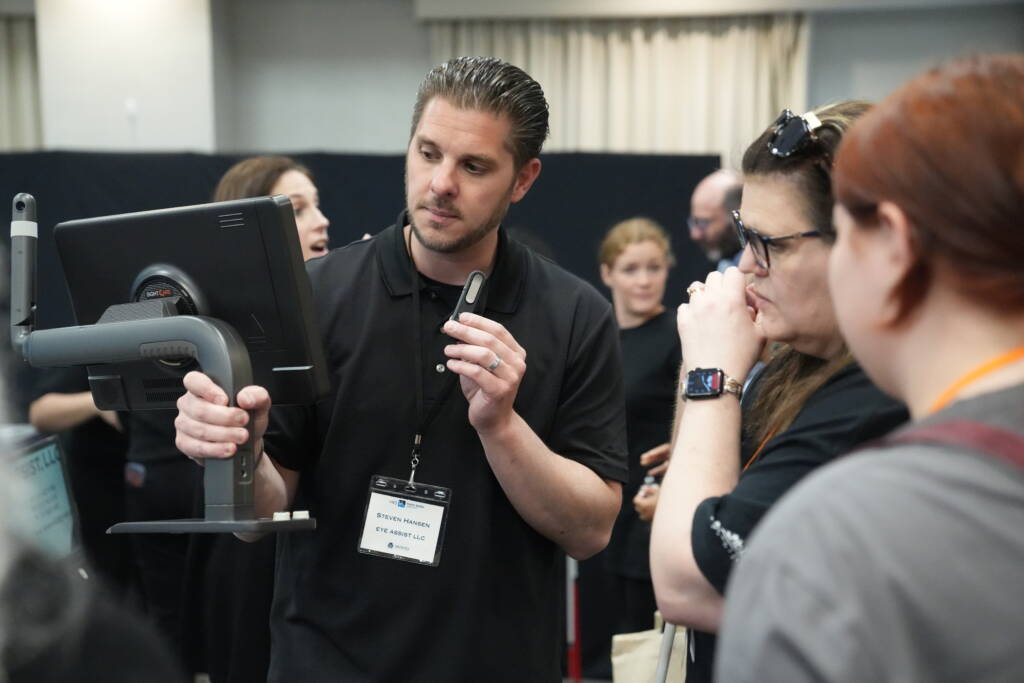 A vendor representative speaks into a small hand-held hearing aid microphone while showing attendees a display monitor.