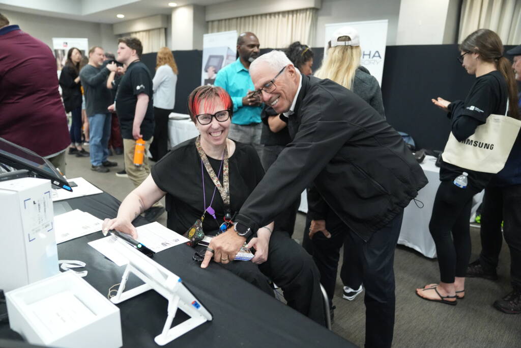 Jamie engages with a vendor at a table featuring magnifying and other assistive technology devices.