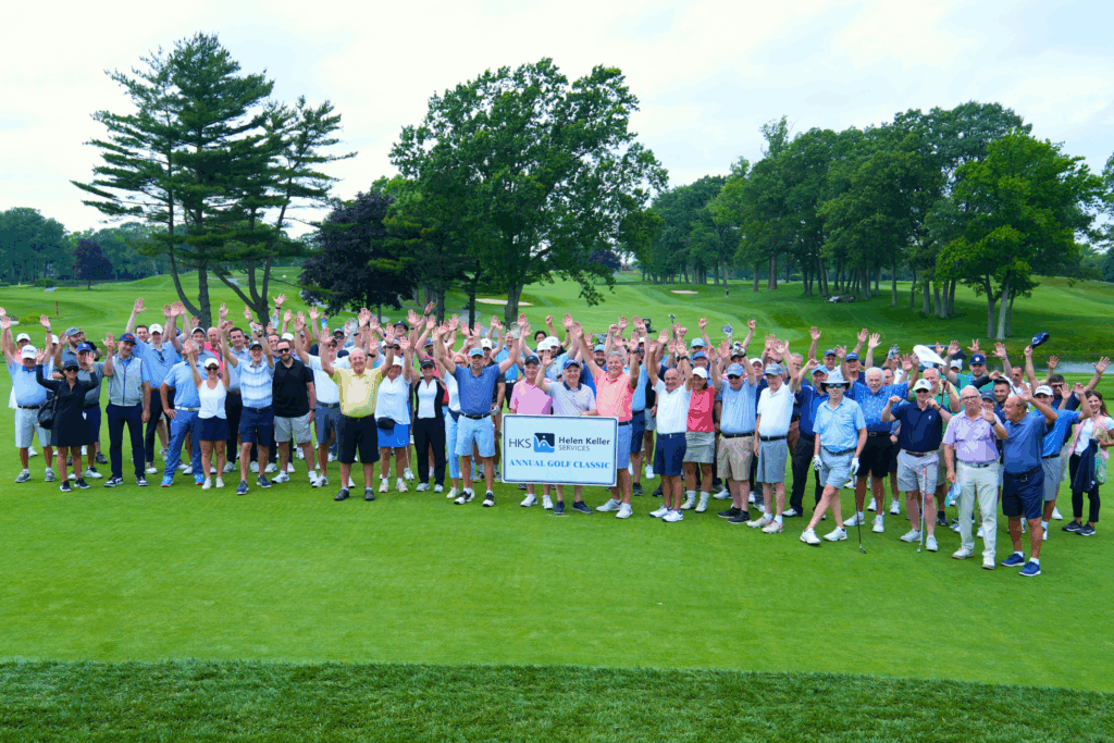 Group photo of Helen Keller Services Golf Classic participants standing together on a large practice green at a golf course.