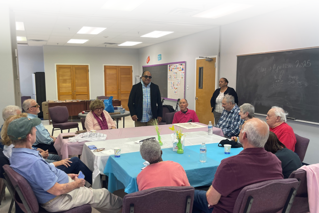 Seniors gathered around tables with blue and pink tablecloths at the Hempstead Senior Center, engaged in discussion with standing presenters, one wearing Meta Ray Ban sunglasses.