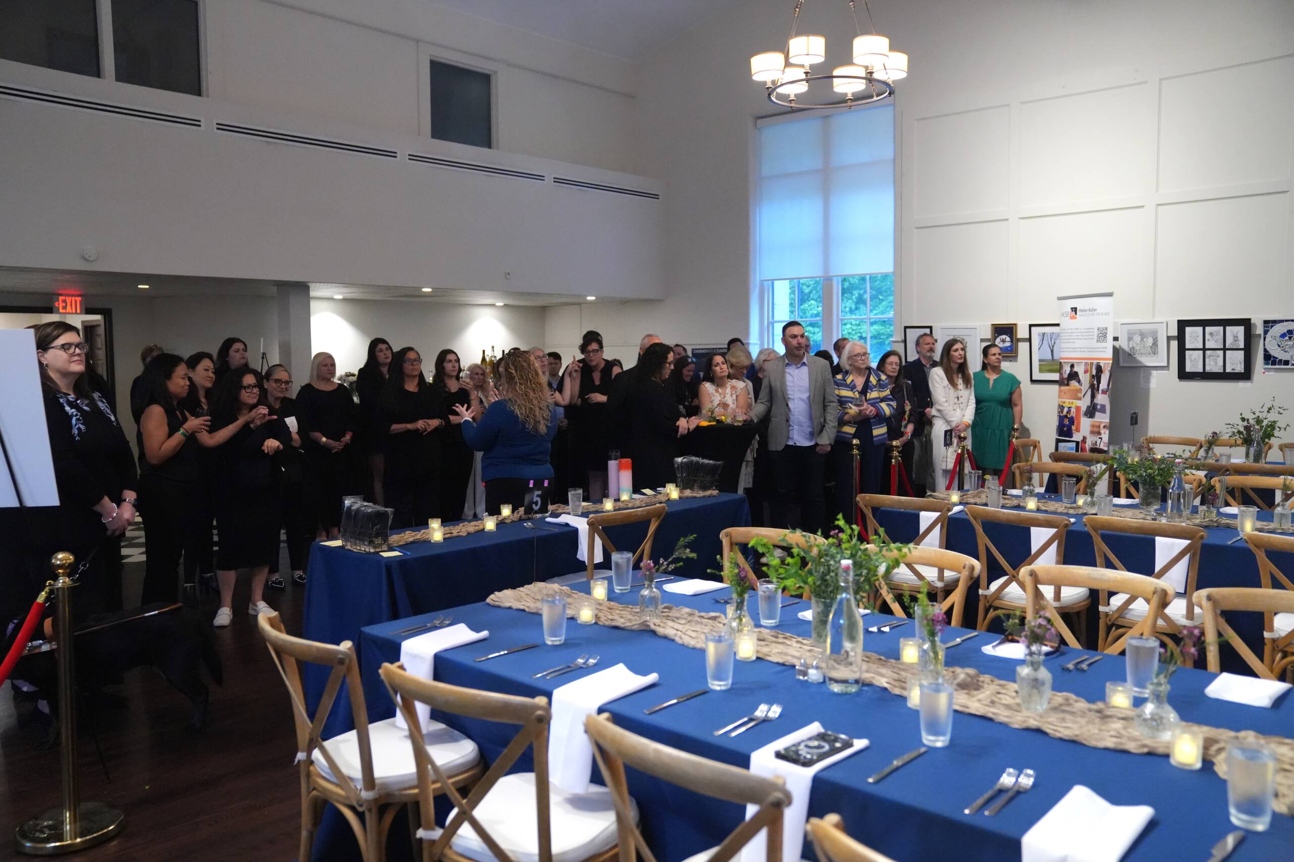 A large group of guests are standing together behind set tables  looking towards a speaker who is not in the picture.   